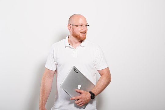 Strahlender Mann mit rötlichem Bart und Brille und weißem Poloshirt hält einen Mac in der Hand