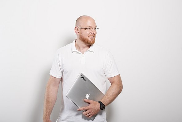 Strahlender Mann mit rötlichem Bart und Brille und weißem Poloshirt hält einen Mac in der Hand
